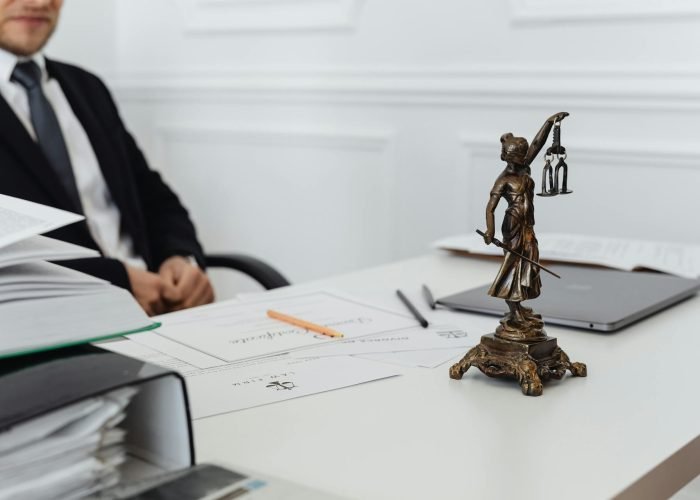 Estátua da Justiça (Themis) de bronze em cima de uma mesa de escritório branca com documentos jurídicos, caneta e um notebook. Ao fundo, um advogado de terno e gravata está sentado.